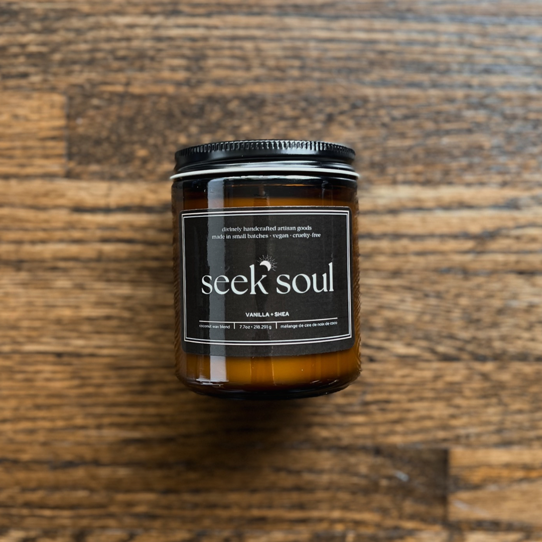A photo of our Vanilla + Shea Coconut Candle. It is in a glass amber jar with a black metal lid. There is a black rectangular label on the front with the Seek Soul logo on it. It has a cotton wick and the wax is an off-white colour.