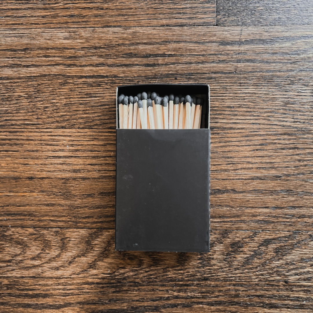 Photo of a black box of our wooden matches. The matches are a light coloured wood with black tips. Sticks are approximately 3” tall. Box has a strike strip on the side.