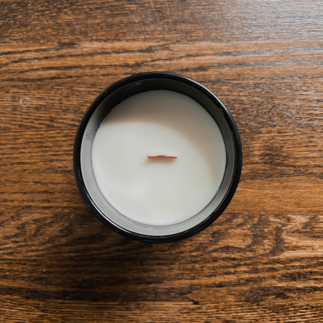 Photo of our Vanilla + Shea Hemp Candle. It is in a matte black glass jar, with no label on it. The wax is white and the candle has a wooden wick. It is just over 4” tall.