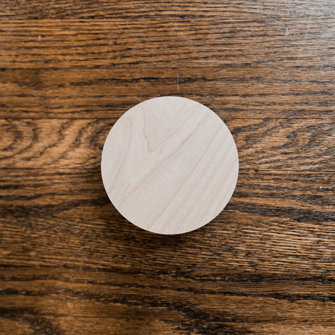 A photo of our Maple Wood Lid. It’s circular and has a diameter that is approximately 3.75” wide. It has a unique grain pattern and light beige colour.