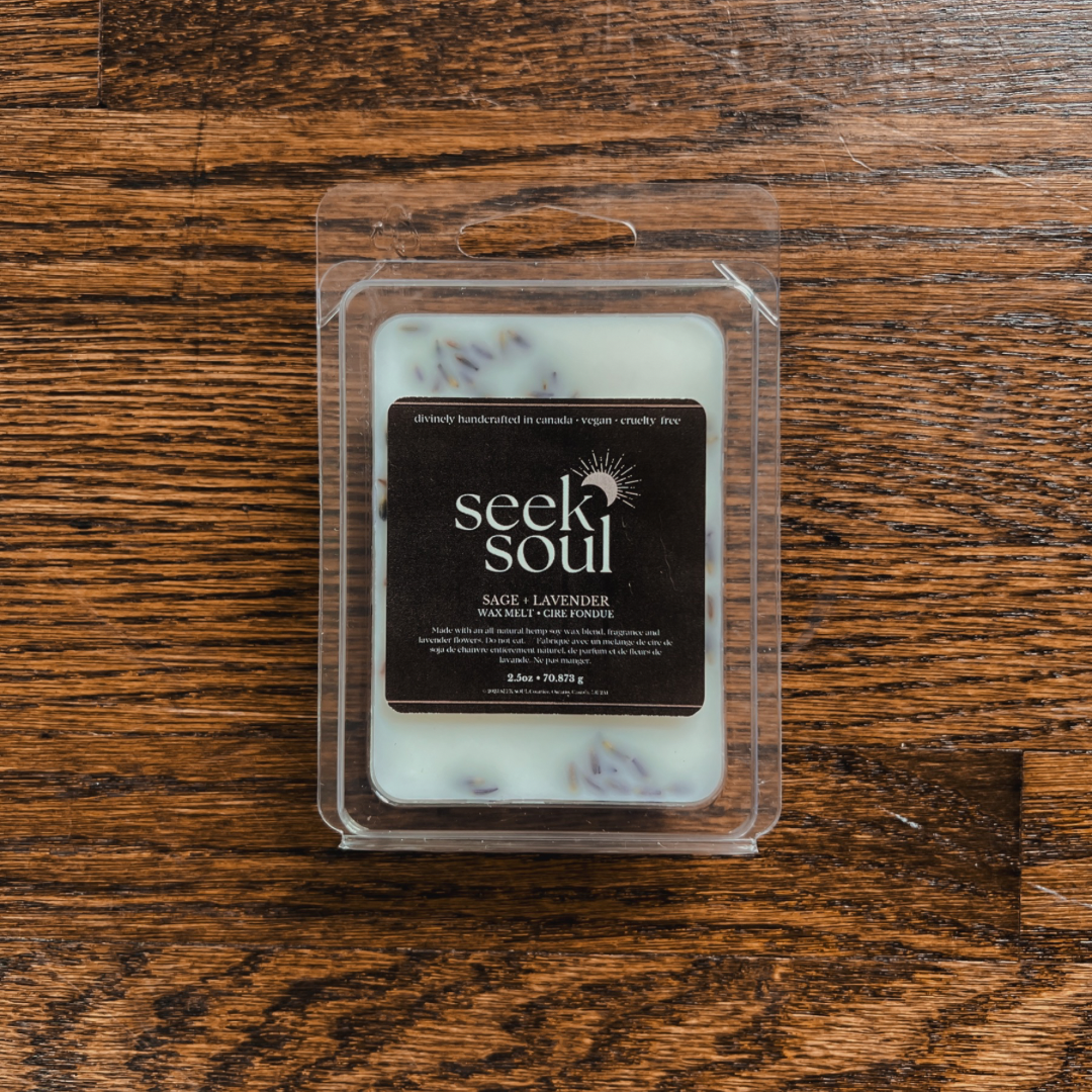 Photo of our Sage + Lavender Wax Melts. Melts are white in colour with purple lavender buds scattered throughout. There are 6 large cubes. Packaging is clear, recyclable and approximately 4” long. Box has a black label on the front with the SEEK SOUL logo on it.