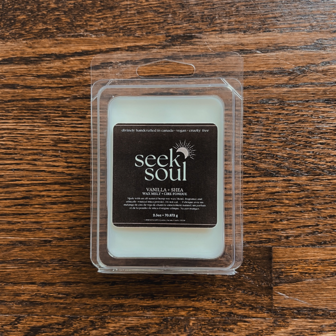 Photo of our Vanilla + Shea Wax Melts. Melts are off-white in colour with a subtle gold sparkle to them. There are 6 large cubes. Packaging is clear, recyclable and approximately 4” long. Box has a black label on the front with the SEEK SOUL logo on it.
